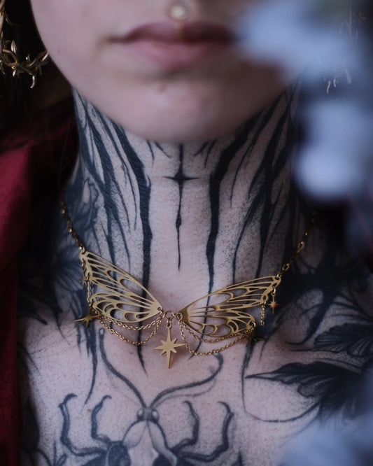 Collier Éos en acier inoxydable doré avec ailes ajourées, fines chaînettes et étoiles suspendues, porté en ras du cou.