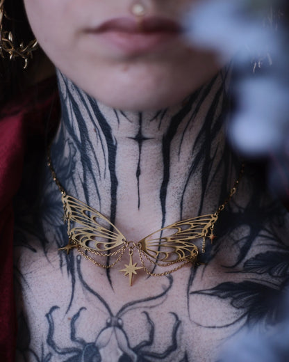 Collier Éos en acier inoxydable doré avec ailes ajourées, fines chaînettes et étoiles suspendues, porté en ras du cou.