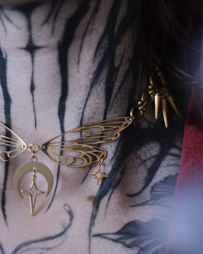 Collier Nyx en acier inoxydable doré avec ailes ajourées, breloques lune et étoiles, porté autour du cou.