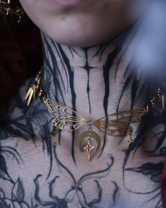 Collier Nyx en acier inoxydable doré avec ailes ajourées, breloques lune et étoiles, porté autour du cou.