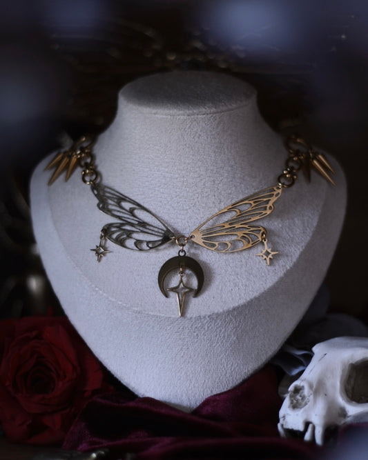 Collier Nyx en acier inoxydable doré avec ailes ajourées, breloques lune et étoiles, porté autour du cou.